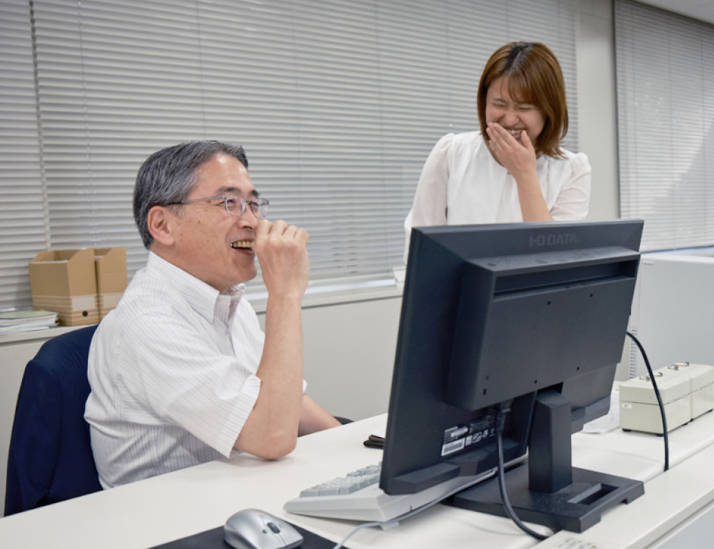 上司の机のところで、女性社員が楽しそうに笑っている様子。話題は近くのおすすめランチの話らしい。